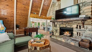 A spacious living area with a stone fireplace, a flat-screen TV mounted above it, and a cozy seating area with a green sofa and a wooden coffee table. - Sugar Bear Cabin- 4bdrm, Semi-Private, Fire-pit, Hot Tub, and Tranquil setting (Pigeon Forge)