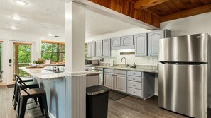 A modern kitchen with grey cabinets, a stainless steel refrigerator, and a bar area with stools. - Sugar Bear Cabin- 4bdrm, Semi-Private, Fire-pit, Hot Tub, and Tranquil setting (Pigeon Forge)