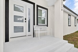 A white house with a front door, two windows, and a bench outside. - Elegant New 3BR Home • Modern Finishes & Natural Light (Thonotosassa)