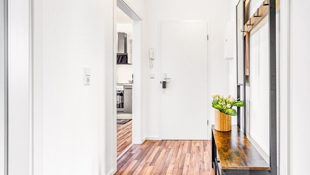 Un pasillo con suelo de madera, paredes blancas, una puerta, un banco con una planta y una ventana con persianas.