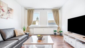 Una sala de estar moderna con un sofá de cuero, una mesa de centro con un tazón, un televisor y vistas a una torre a través de amplios ventanales.