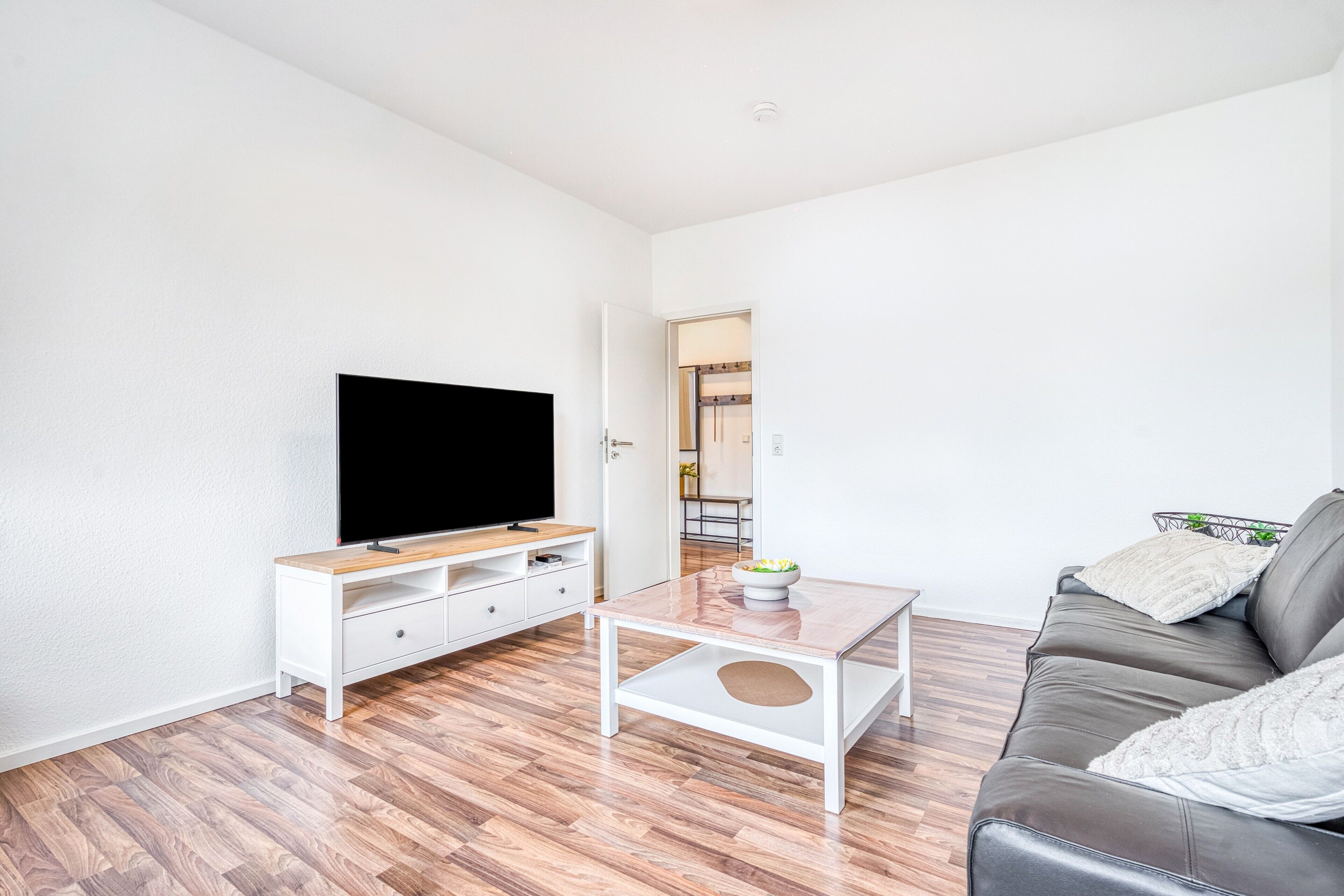 A modern living room with a flat-screen TV, a white coffee table, a leather sofa, and a wooden floor.