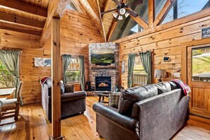A cozy cabin interior with wooden walls, a stone fireplace, leather sofas, and a dining area. - 1/2 Mile Parkway*2BR*Hot Tub*Games*Families*Pools (Pigeon Forge)