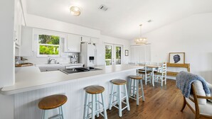 A modern kitchen with white cabinetry, a large island with stools, and a dining area with wooden chairs and a table. - WELCOME TO DOC'S HOLIDAY! (St. George Island)