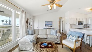 A spacious living room with a large window, a ceiling fan, a sofa, a coffee table, and a kitchen area in the background. - WELCOME TO DOC'S HOLIDAY! (St. George Island)