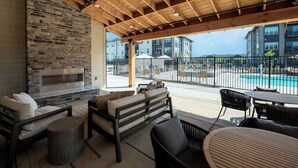 Un espace extérieur aménagé, abrité, avec une cheminée en pierre, du mobilier de jardin et une vue sur une piscine et des bâtiments en arrière-plan.