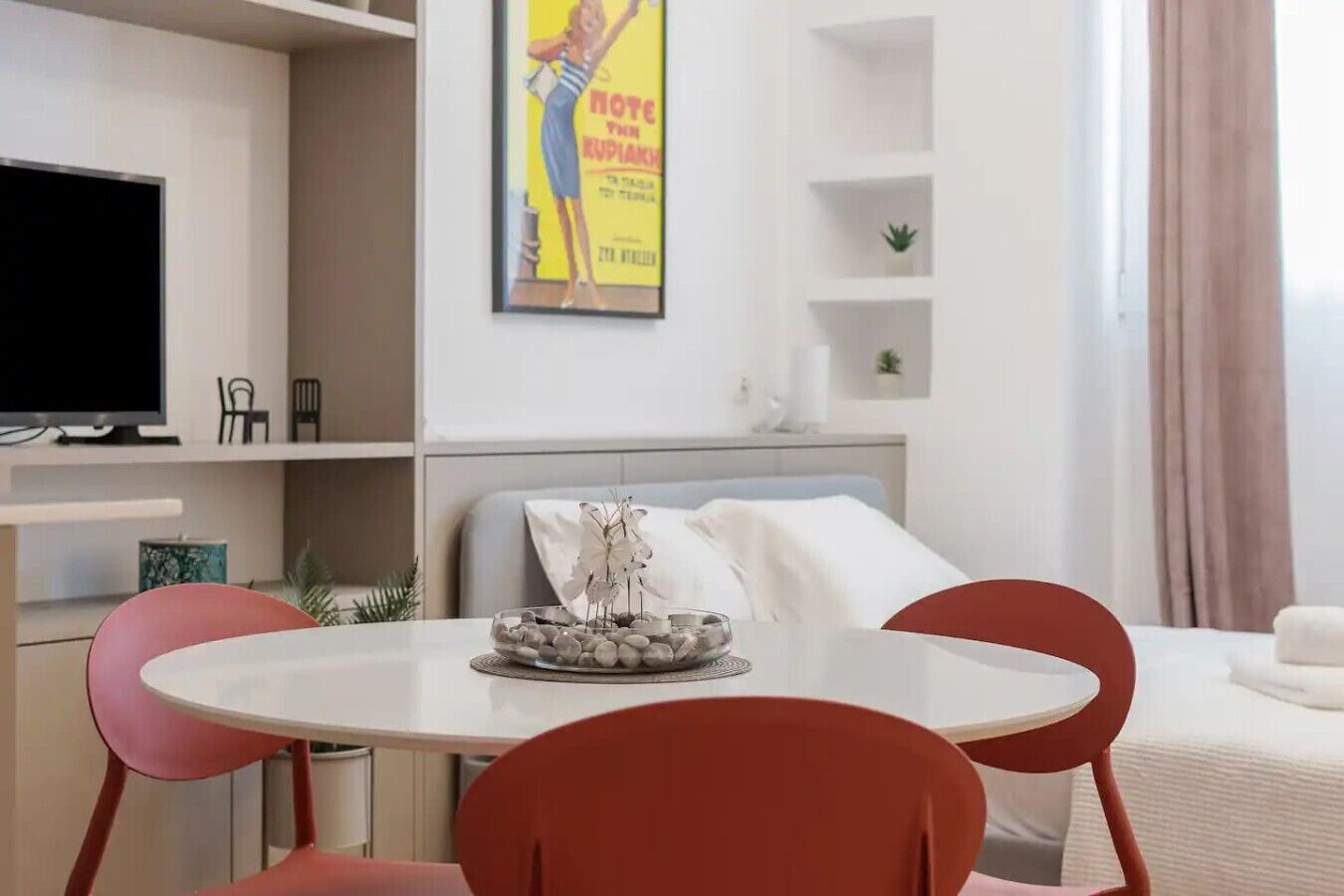 A modern hotel room with a bed, a dining table with chairs, a TV, and a poster on the wall.