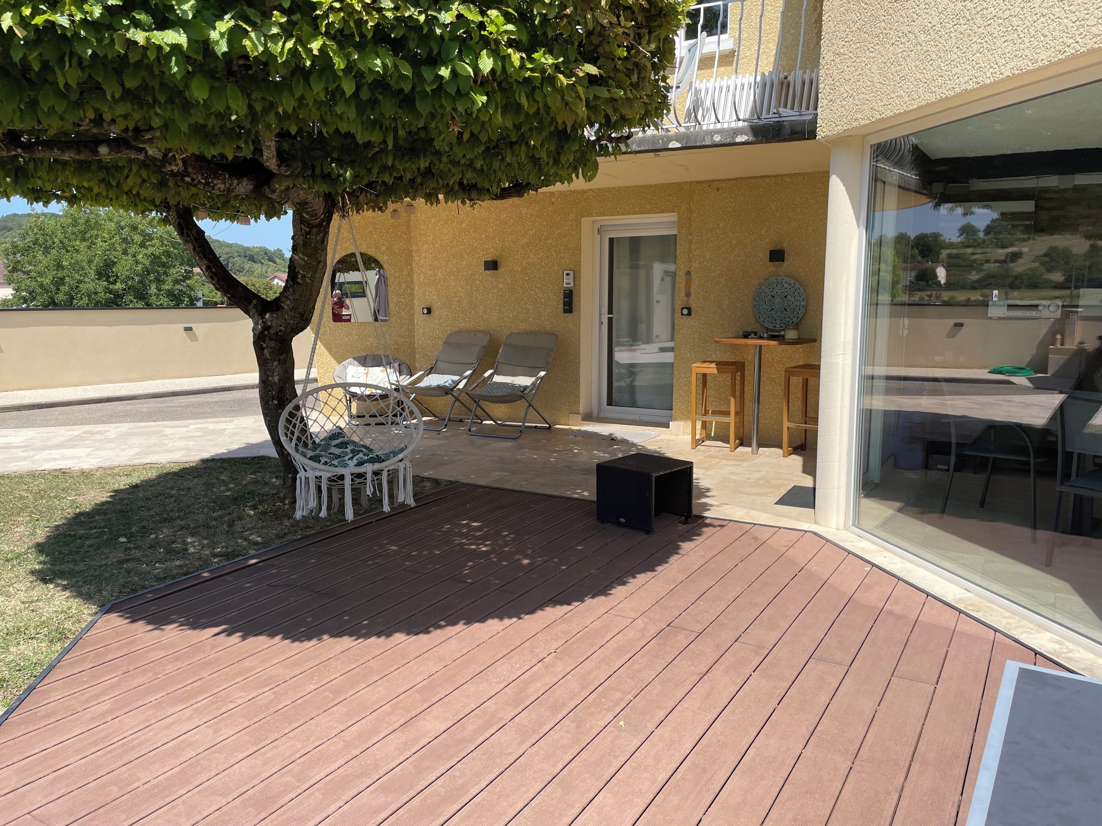 A covered outdoor area with a wooden deck, a hanging chair, two lounge chairs, a small table, and a glass door leading to a patio.