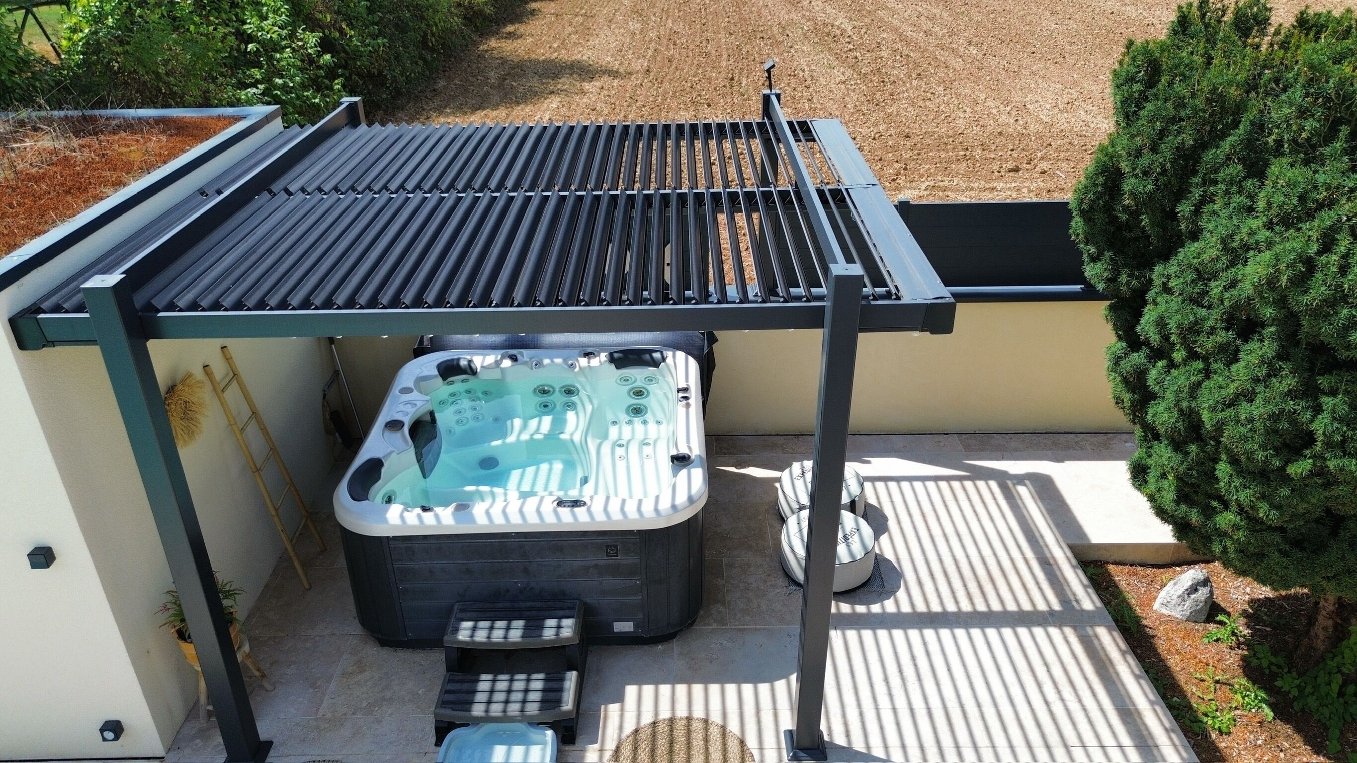 Un bain à remous installé sous une pergola dotée d’un toit en métal, entouré d’un espace patio avec une petite table et un balai.