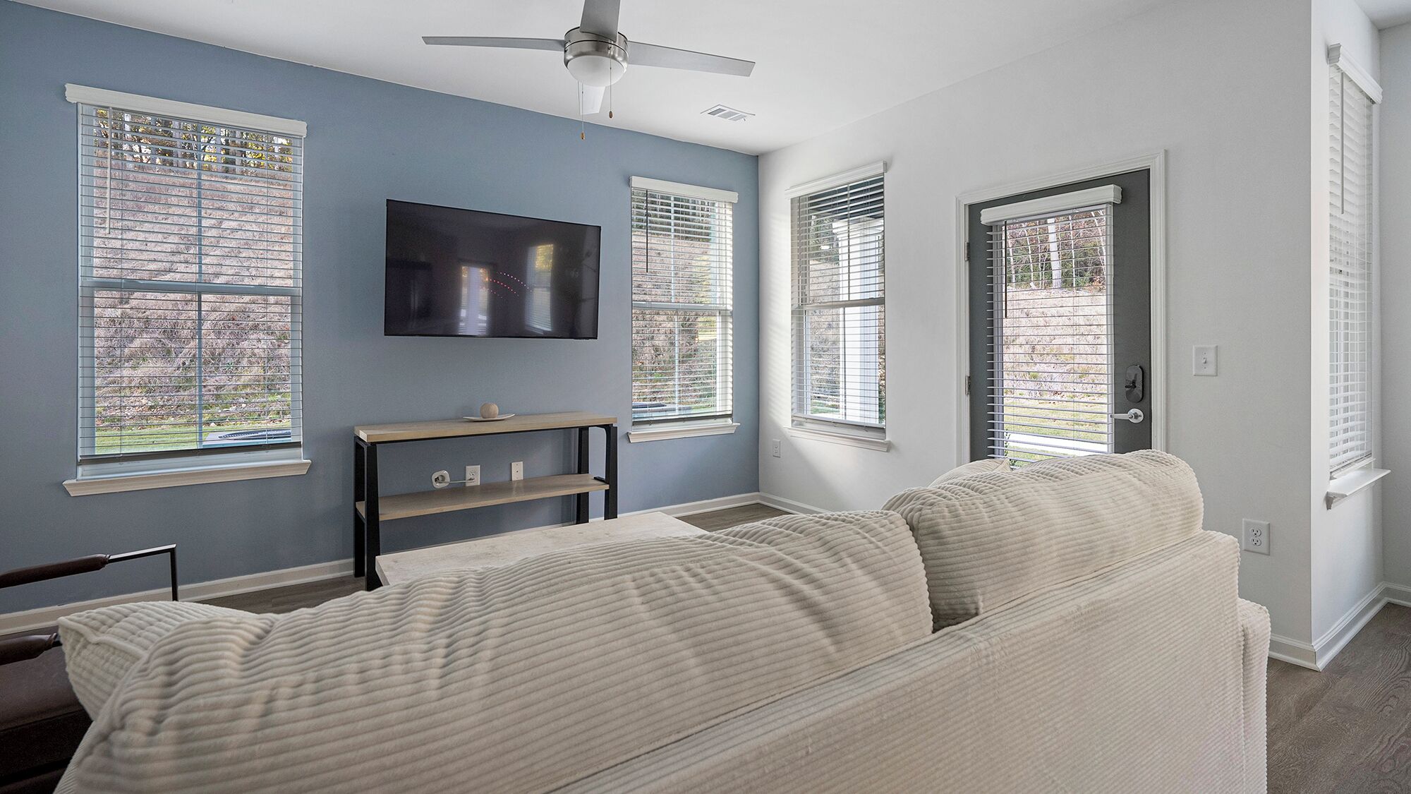A living room with a sofa, a TV mounted on the wall, and windows with blinds.
