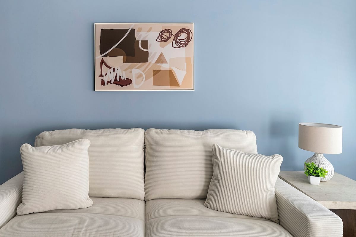 A light-colored sofa with two pillows against a blue wall, a framed abstract artwork above it, and a table with a lamp and a plant to the right.