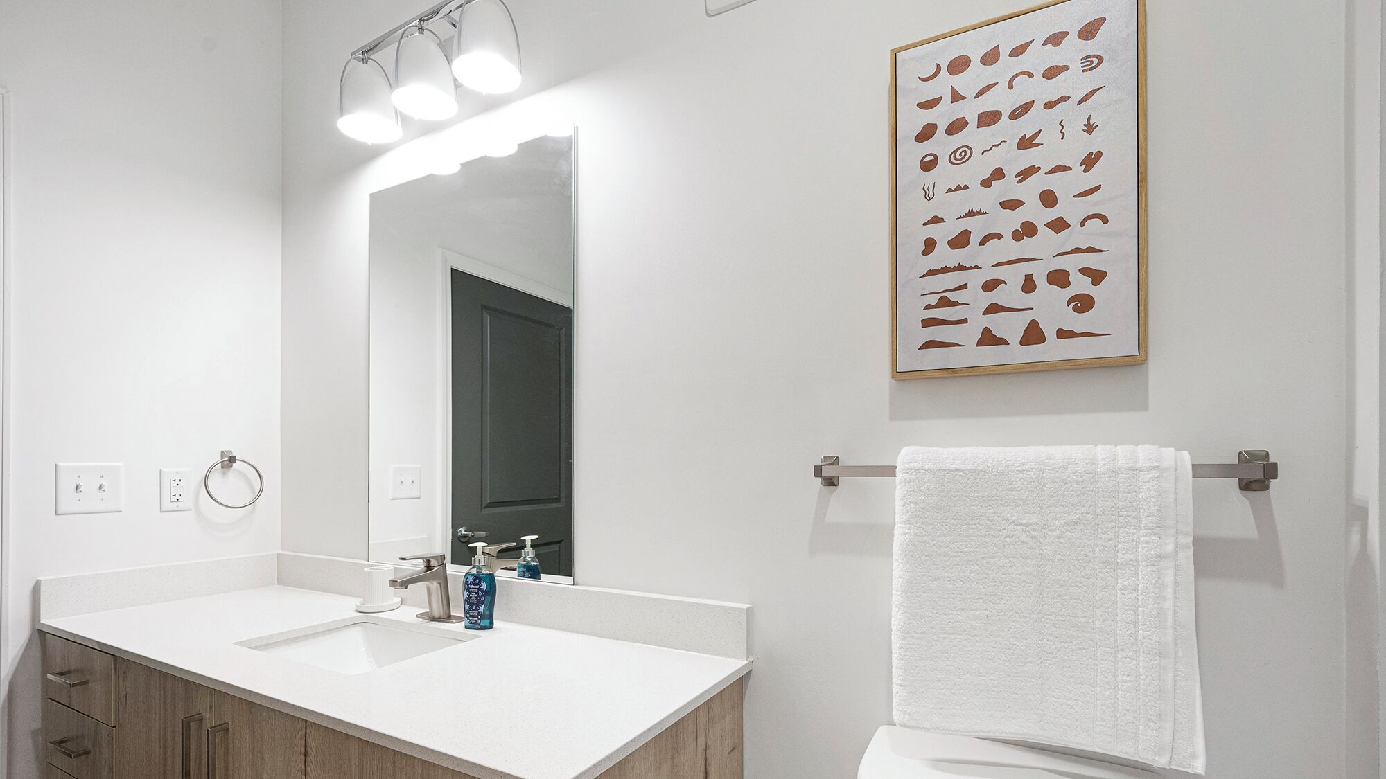 Ein Badezimmer mit einem weißen Waschbecken, einem Spiegel, einem Handtuchhalter und einem gerahmten Kunstwerk an der Wand.