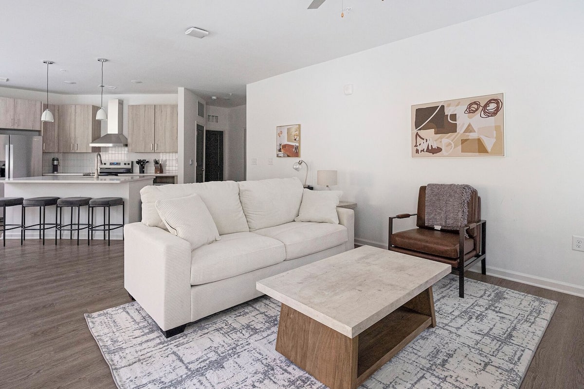A modern living room with a sofa, coffee table, and a chair. There is a kitchen area with a dining set and a bar.