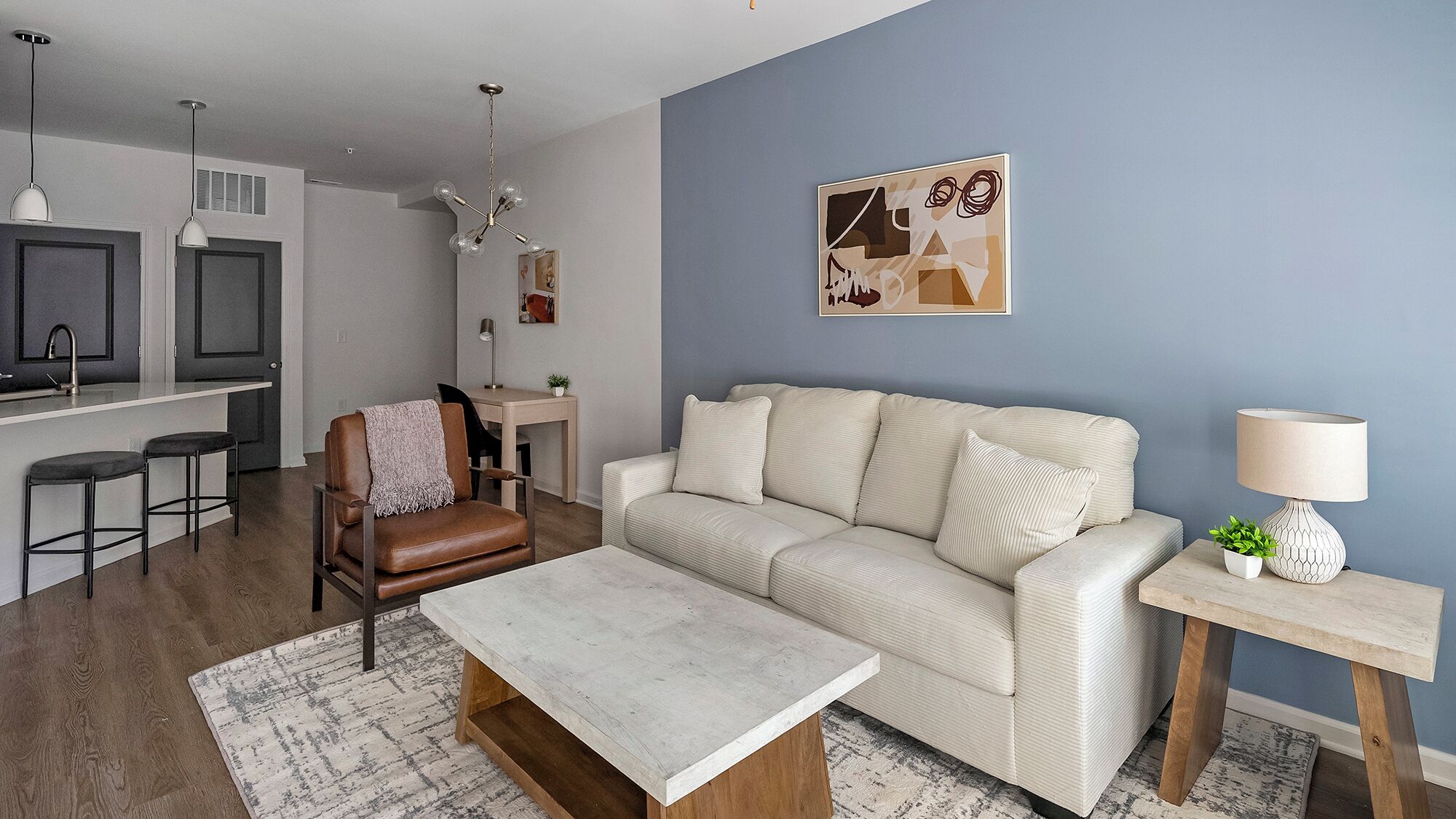Een moderne woonkamer met een bank, salontafel en barstoelen. Er is een eethoek met een tafel en stoelen, en een keuken met een aanrecht en kasten.
