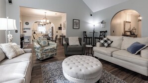 Amplia sala de estar con sofá, sillón, reposapiés y una zona de comedor al fondo.