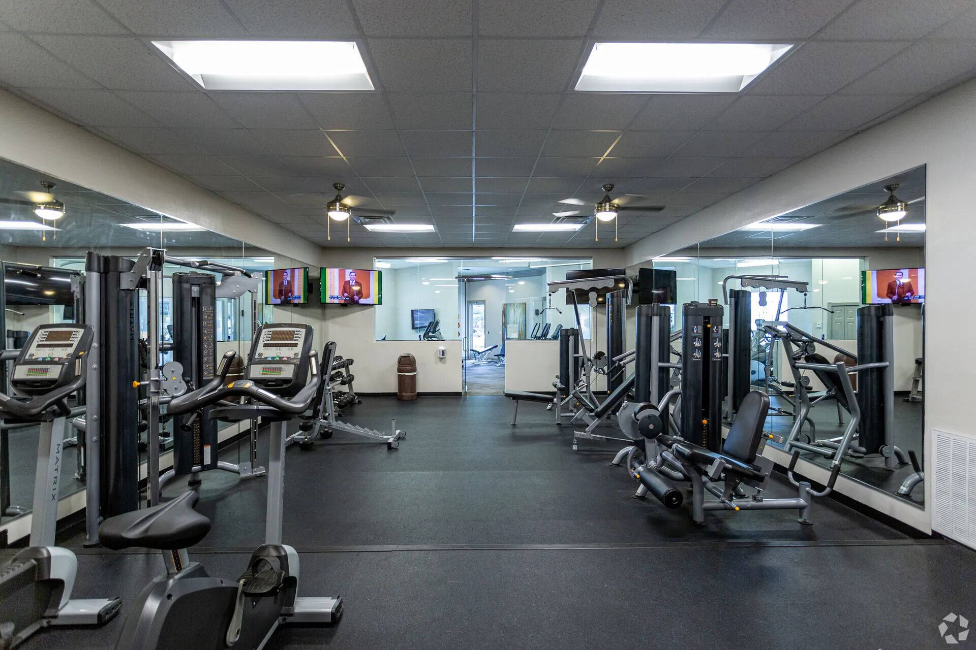 A gym with various exercise machines, mirrors, and ceiling fans.
