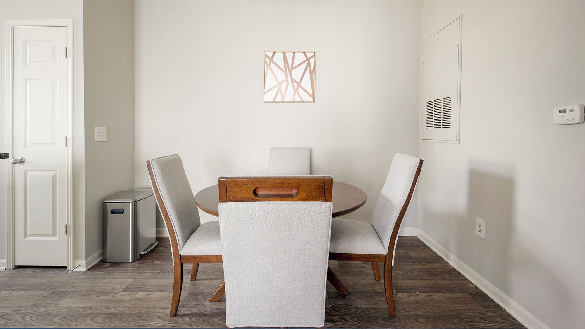 Un coin repas comprenant une table ronde, quatre chaises, une petite poubelle et une bouche d’aération fixée au mur.
