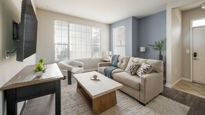 A modern living room with a beige sofa, a wooden coffee table, and a TV mounted on the wall. - Landing Furnished Apartments near Barfield (Murfreesboro)