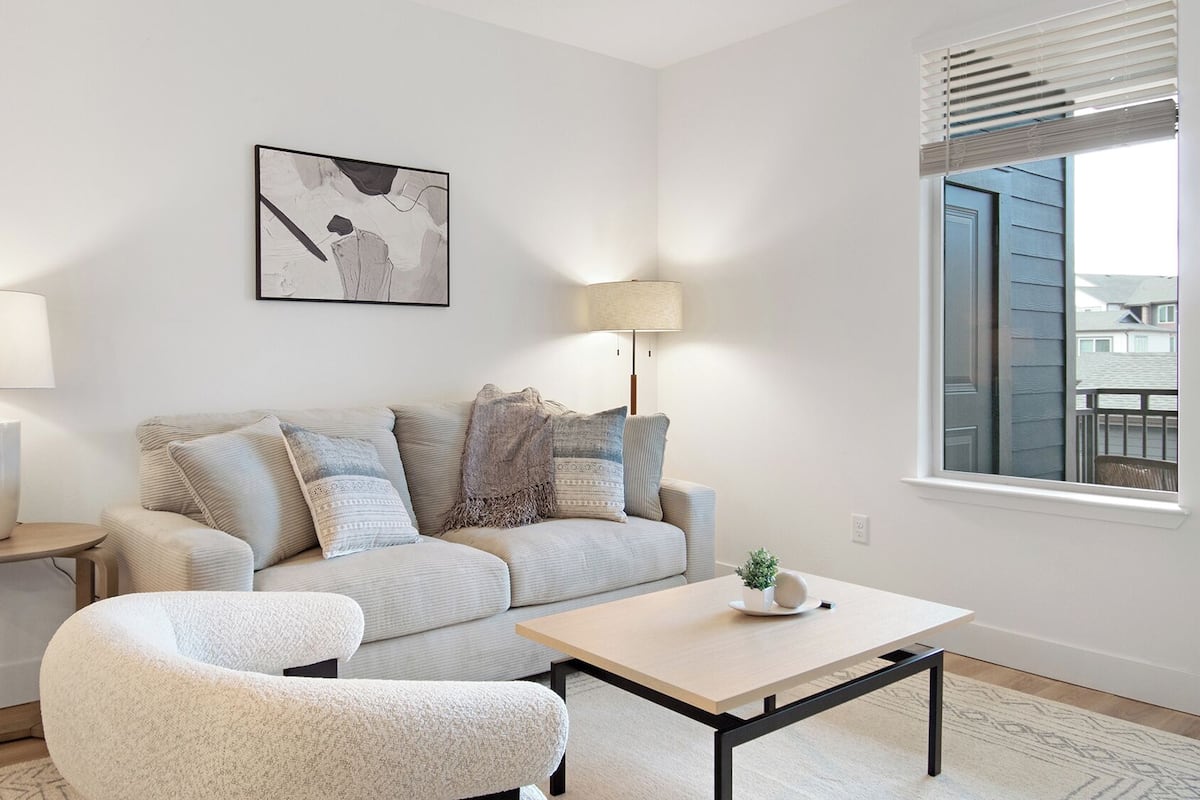 A modern living room with a sofa, armchair, and coffee table. There are two lamps, a painting on the wall, and a window with blinds.