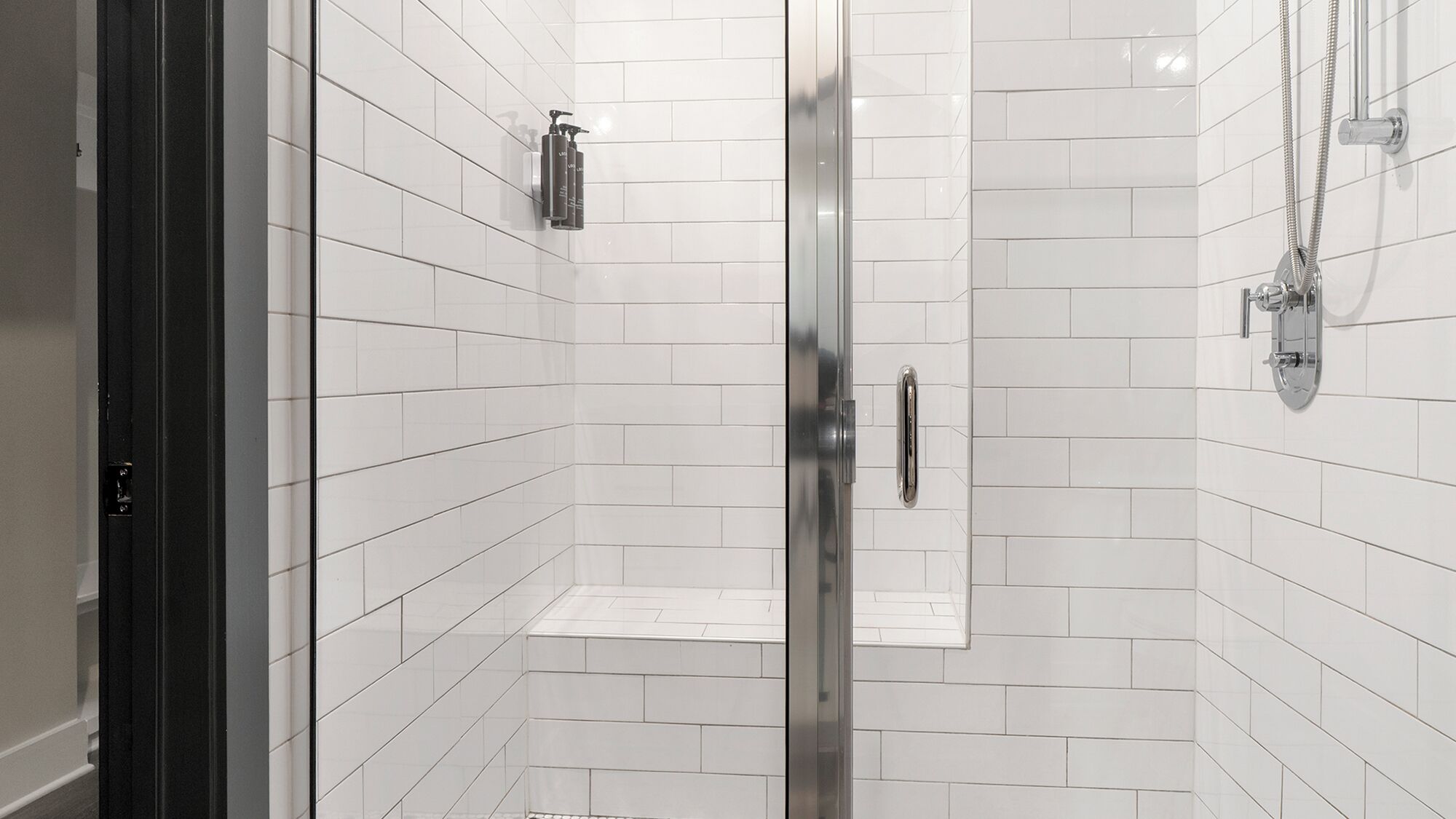 A modern shower with white tiles, a glass door, and a handheld showerhead.
