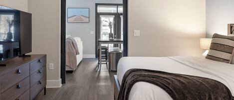 A hotel room with a bed, a TV on a dresser, a desk with a chair, and a window with blinds.