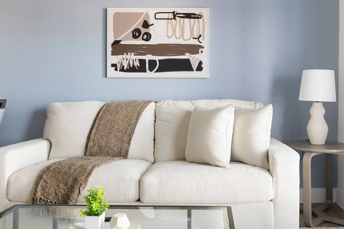 Un salon avec un canapé blanc, une table basse en verre, une lampe et une œuvre d’art murale.