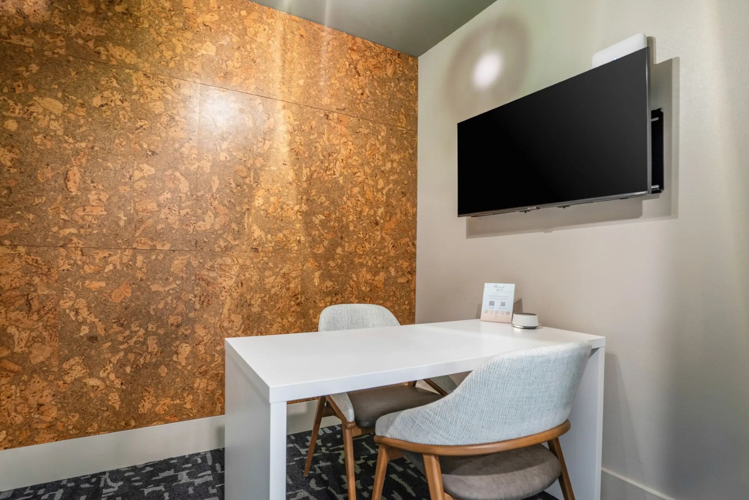 A modern room with a white table and two chairs, a cork wall, and a mounted TV.