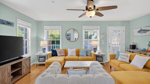 A living room with a yellow sofa, a white armchair, a TV, and a ceiling fan. - Sandy Toes Hideaway 3 Bedroom Home (Tybee Island)