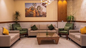 A lobby with a beige sofa set, green armchairs, a wooden coffee table, and a large painting of a building.