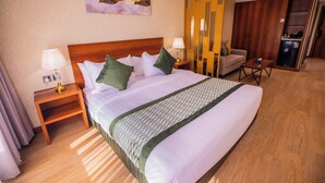 A hotel room with a large bed, two bedside tables with lamps, a sofa, a small table, a refrigerator, and a decorative wall piece.
