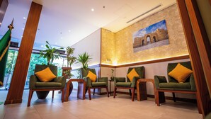 A lobby with four green armchairs with yellow cushions, a large painting of an archway, and a potted plant.
