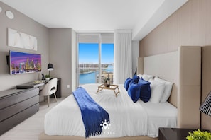A modern hotel room with a large bed, a desk, a TV, and a view of a cityscape. - Luxury 2BR Apt w/ Stunning Views | Amazing Pool (Hollywood)