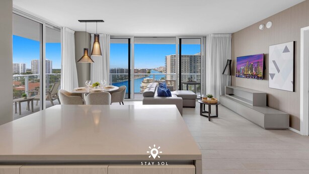 A modern hotel room with a large dining table, a sofa, and a view of a cityscape and water. - Luxury 2BR Apt w/ Stunning Views | Amazing Pool (Hollywood)