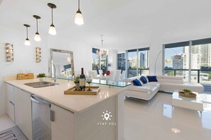 A modern kitchen with a central island, a dining area with a glass table, and a living room with a white sofa and large windows offering city views. - Apt w/ Private Balcony + Incredible Pool (Hallandale Beach)