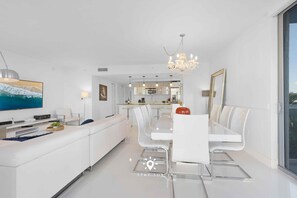 A modern living room with a white sofa, a dining table with chairs, a chandelier, and a large TV. - Apt w/ Private Balcony + Incredible Pool (Hallandale Beach)