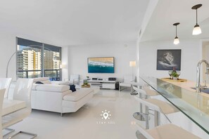 A modern living room with a white sofa, a glass coffee table, and a large window offering a city view. - Apt w/ Private Balcony + Incredible Pool (Hallandale Beach)