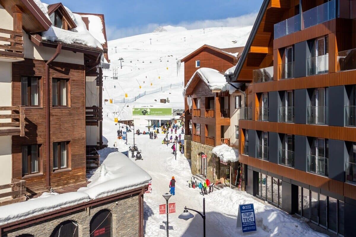 A snow-covered street with buildings on either side, a ski lift in the background, and people walking.