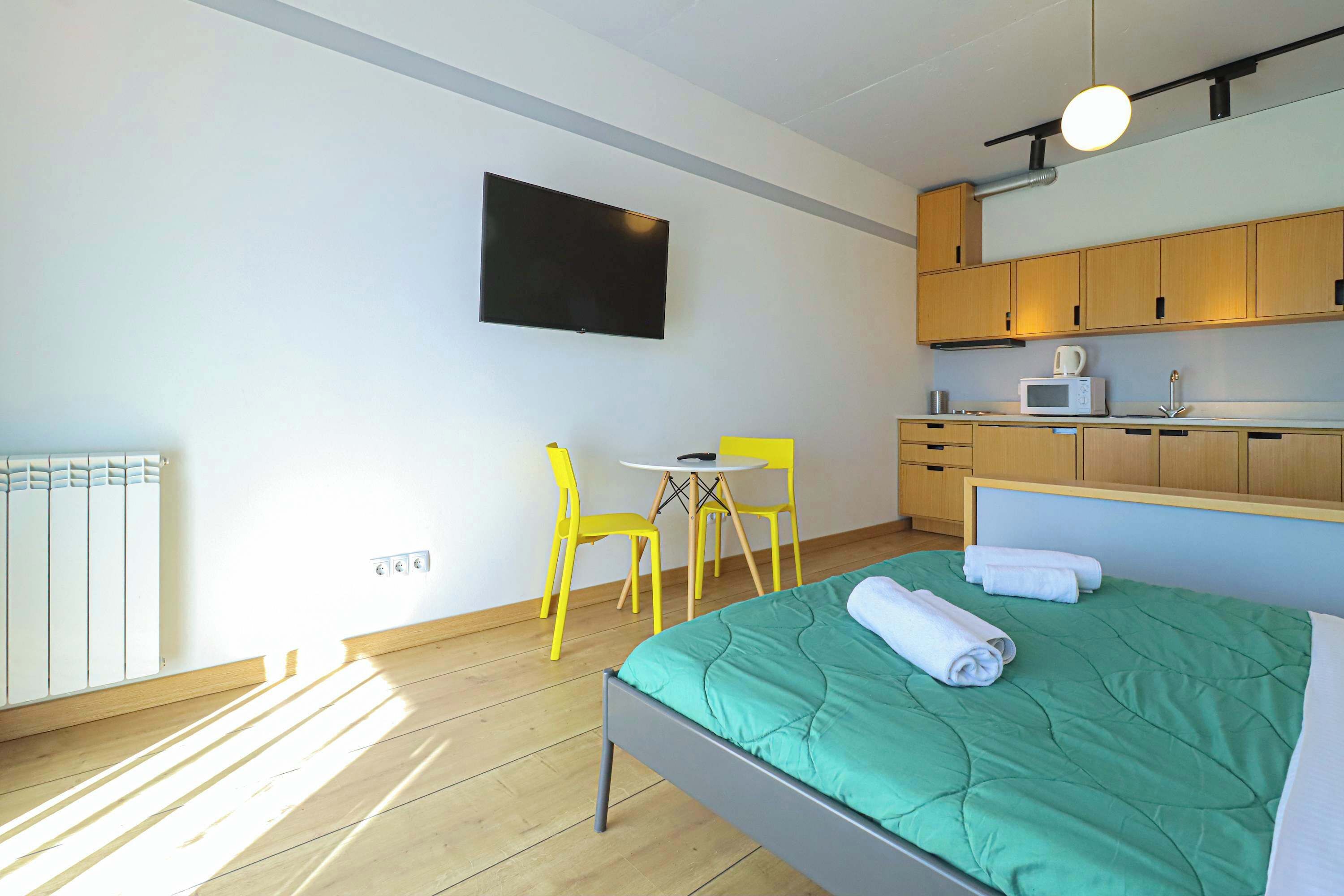 A modern hotel room with a bed, a small table, a chair, a TV, and a kitchenette.