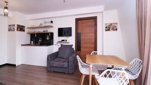 Een kleine woonruimte met een kitchenette, een bank, een eettafel met stoelen en een aan de muur bevestigde televisie.