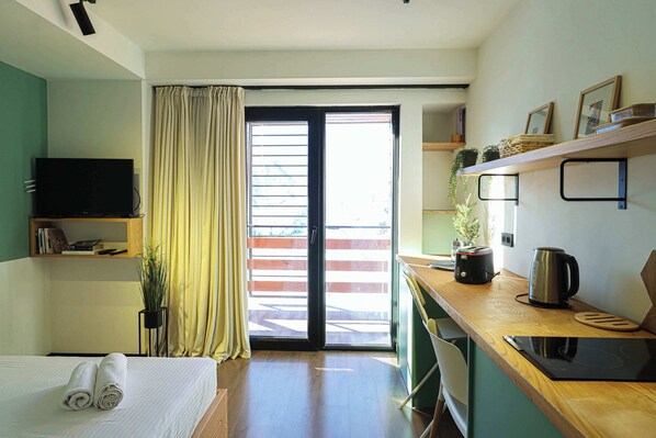 A hotel room with a bed, a TV, a kitchen area with a kettle and toaster, and a sliding glass door to the outside.