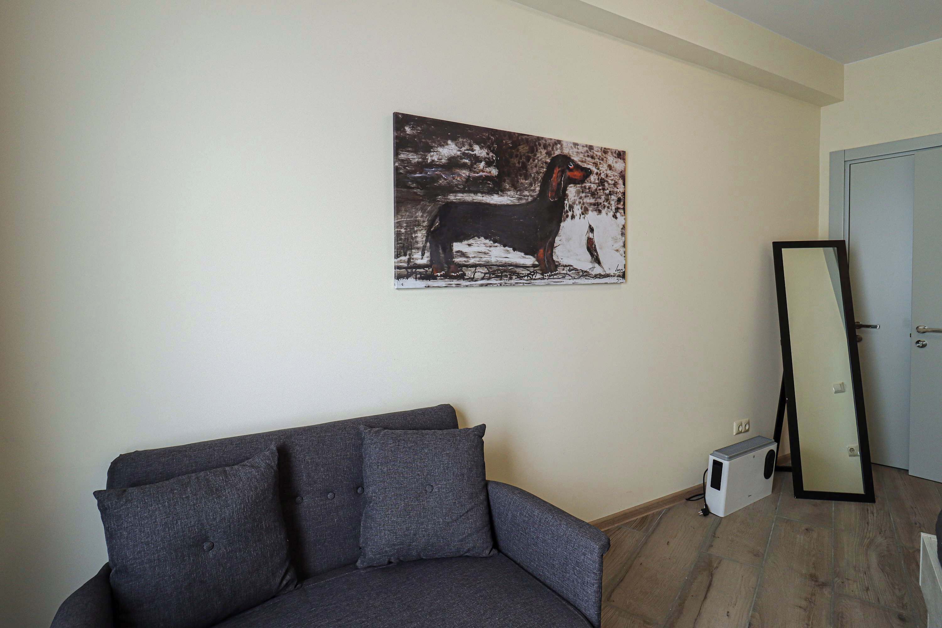 Um quarto com sofá cinzento, um quadro de um cão, um espelho e uma porta branca.