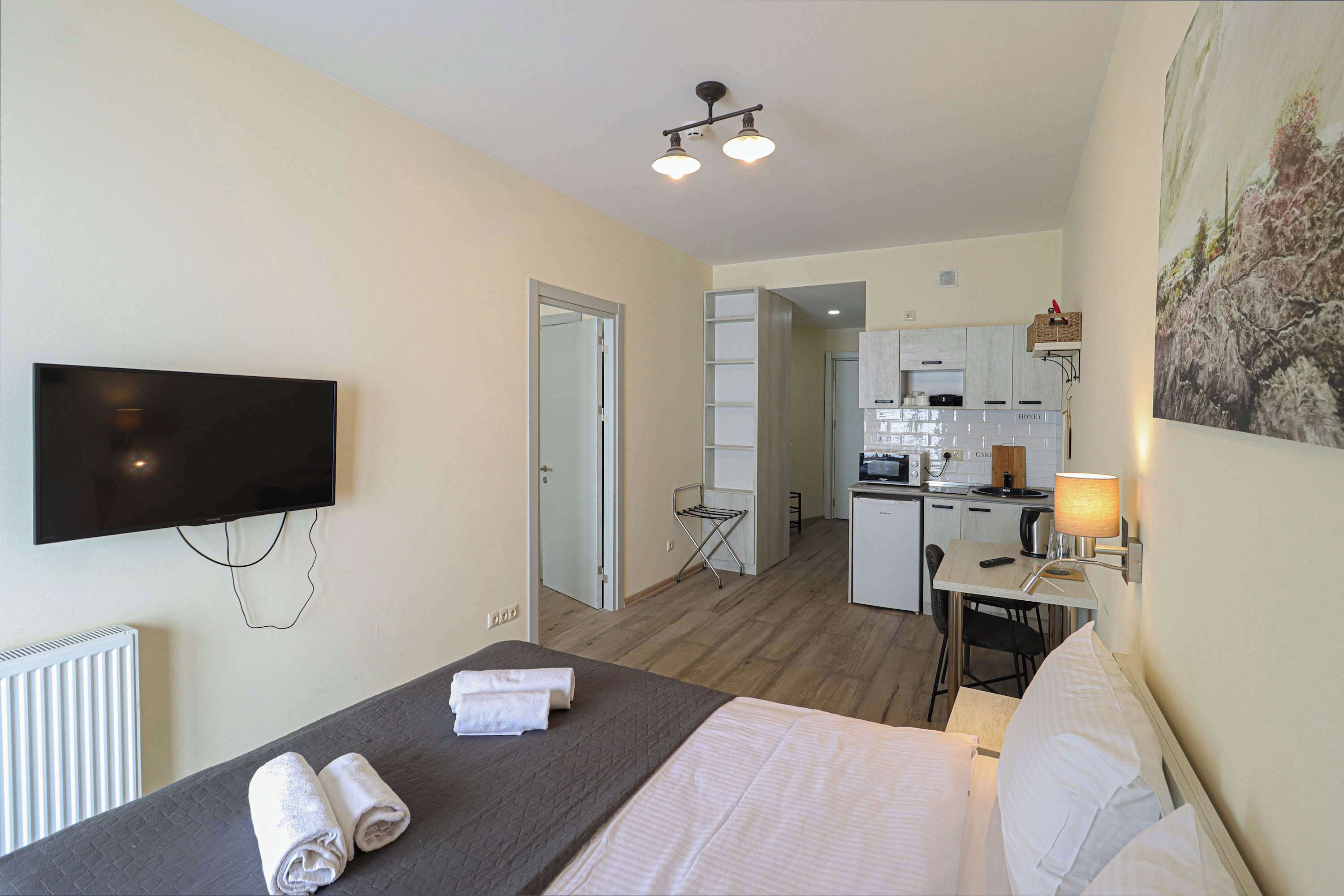 A hotel room with a bed, a TV, a desk with a lamp, and a kitchenette in the background.