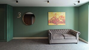 Una hall con una parete verde, un cartello con il numero '3', uno specchio rotondo, un divano trapuntato e un grande quadro astratto.