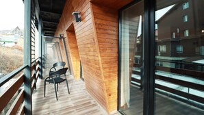 Ein Holzbalkon mit einem Stuhl, einem Metallgeländer und Blick auf ein Gebäude durch eine Glastür.