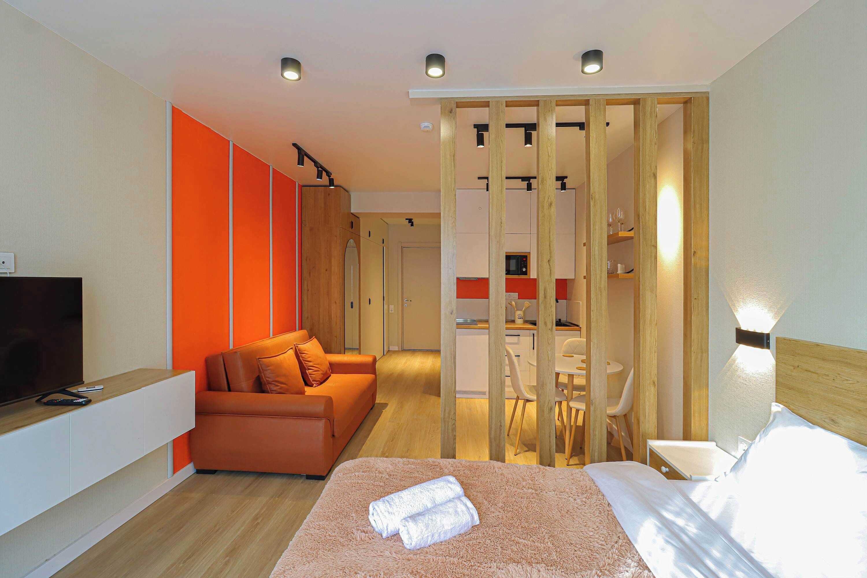 A modern hotel room with a sofa, a bed with a rolled-up towel, a TV, and a kitchenette in the background.