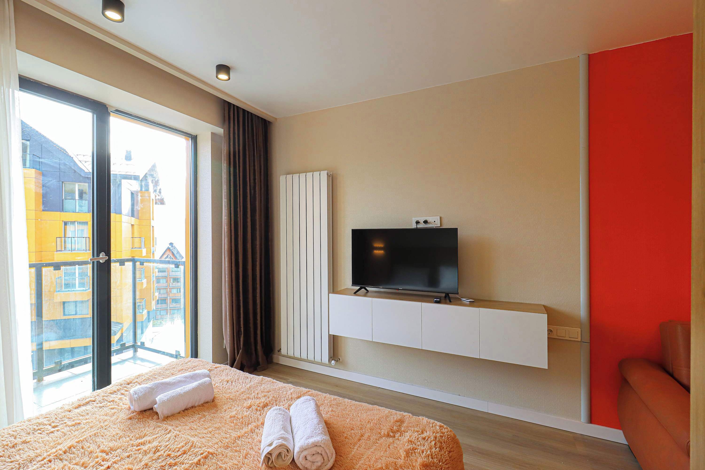 Ein Hotelzimmer mit einem großen Bett, einem an einer weißen Kommode befestigten Fernseher, einem Balkon mit Blick auf Gebäude und einer roten Akzentwand.