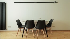Una habitación con piso de madera, pared blanca y radiador negro. Cuatro sillas negras con patas de madera rodean una mesa rectangular.
