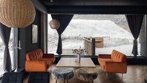 A modern living room with a large window, two orange sofas, a wooden coffee table, and a wicker pendant light. - Marshall Gudauri 2 Room Apt - By Wehost (Gudauri)