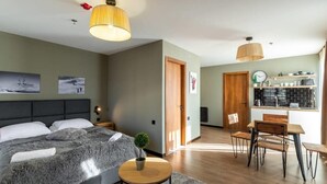 Quarto de hotel moderno com uma cama grande, uma pequena mesa redonda, uma área de refeições com mesa e cadeiras de madeira, e uma cozinha com painel de parede preto e armários de madeira.