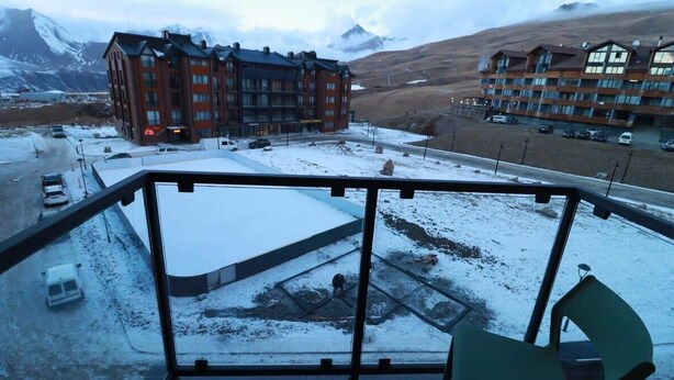 A hotel with a snow-covered parking area and a view of mountains in the background. - Stylish Ski-In/Out Apt- Mountain Views - By Wehost (Gudauri)